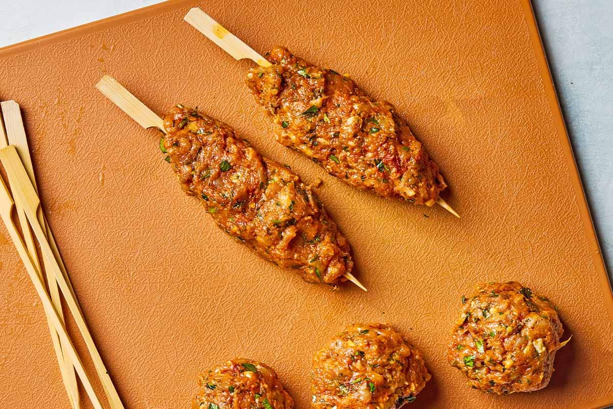 2 uncooked chicken kofta kebabs on a cutting board with bamboo skewers and 3 balls of the kebab mixture.