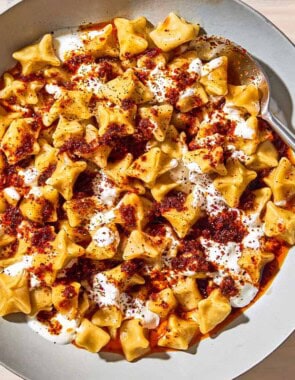 An overhead photo of manti topped with garlic yogurt and spiced oil in a bowl with a spoon. Next to this is a bowl of sumac and a bowl of dried mint.