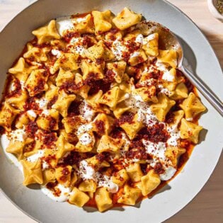 An overhead photo of manti topped with garlic yogurt and spiced oil in a bowl with a spoon. Next to this is a bowl of sumac and a bowl of dried mint.