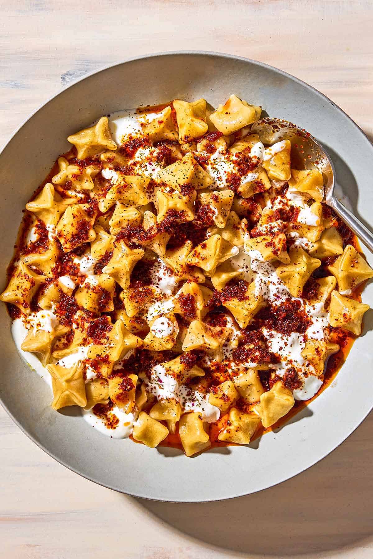 An overhead photo of manti topped with garlic yogurt and spiced oil in a bowl with a spoon.