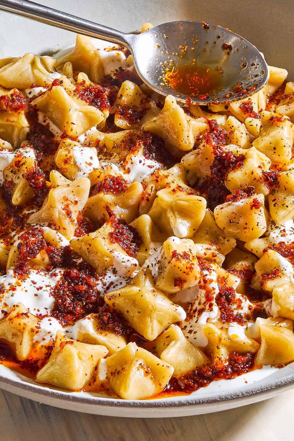 A close up of manti topped with garlic yogurt and spiced oil in a bowl with a spoon.