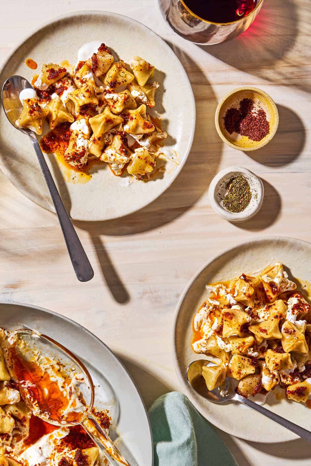 An overhead photo of 2 servings of manti on plates with spoons. Next to this is a glass of red wine, bowls of sumac and dried mint, and the rest of the manti on a serving platter with a spoon.