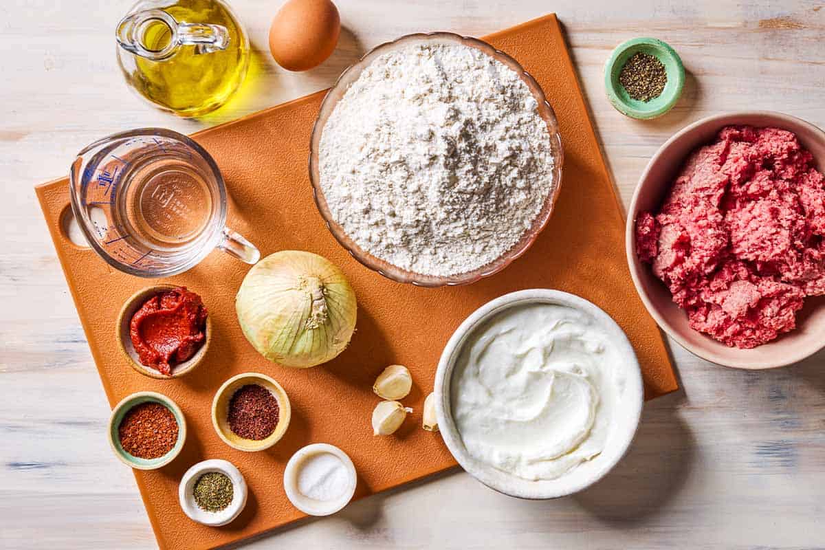 Ingredients for manti including all purpose flour, salt, egg, water, olive oil, ground meat, onion, black pepper, yogurt, garlic, turkish pepper, aleppo pepper, mint, and sumac.