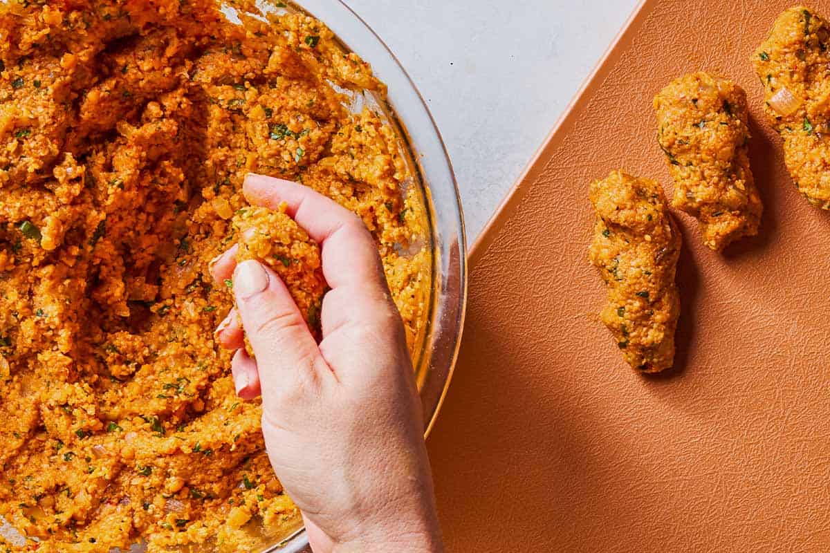 An overhead photo of a hand picking up some of the turkish lentil patty mixture from a bowl. Next to this is a cutting board with 3 uncooked turkish lentil patties on a cutting board.