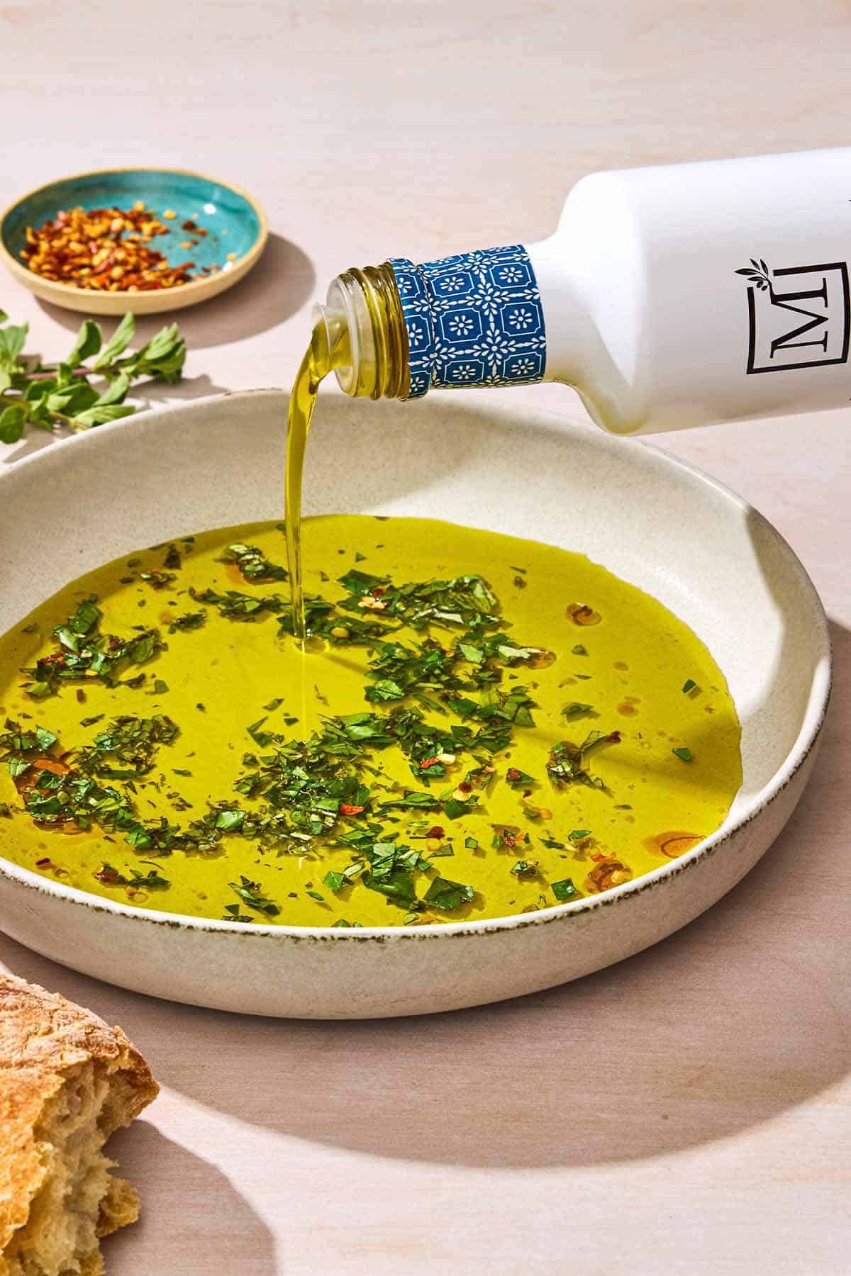 Olive oil being poured from a bottle into a bowl of olive oil seasoned with spices and herbs. Next to this is a small bowl of red pepper flakes.