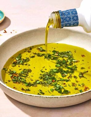 Olive oil being poured from a bottle into a bowl of olive oil seasoned with spices and herbs. Next to this is a piece of crusty bread, a sprig of herbs and a small bowl of red pepper flakes.