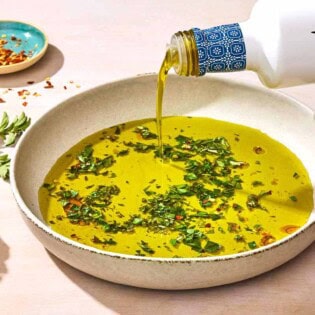 Olive oil being poured from a bottle into a bowl of olive oil seasoned with spices and herbs. Next to this is a piece of crusty bread, a sprig of herbs and a small bowl of red pepper flakes.