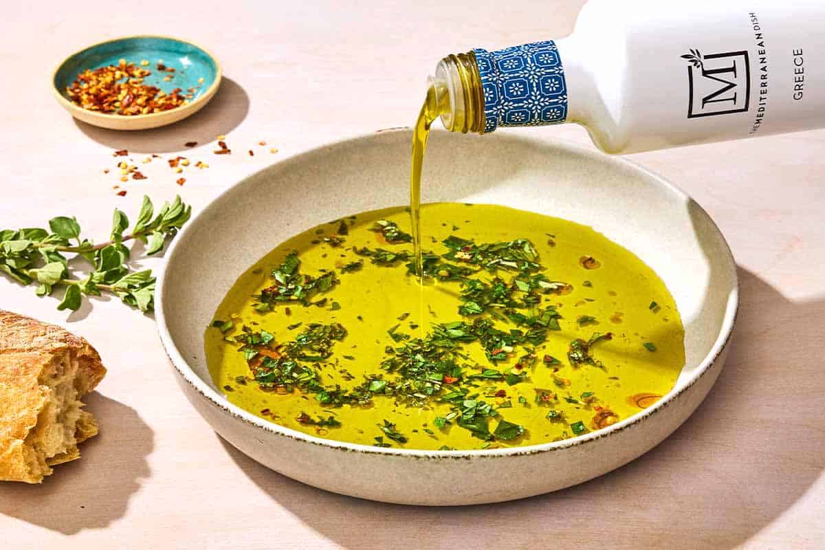 Olive oil being poured from a bottle into a bowl of olive oil seasoned with spices and herbs. Next to this is a piece of crusty bread, a sprig of herbs and a small bowl of red pepper flakes.