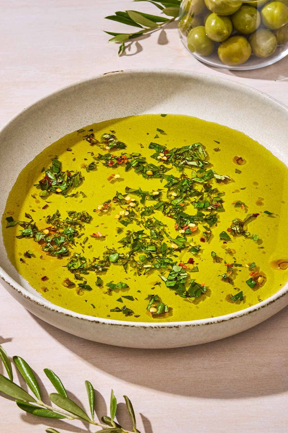 A close up of a bowl of olive oil seasoned with spices and herbs. Next to this is a clear bowl with green olives and sprigs of leaves.