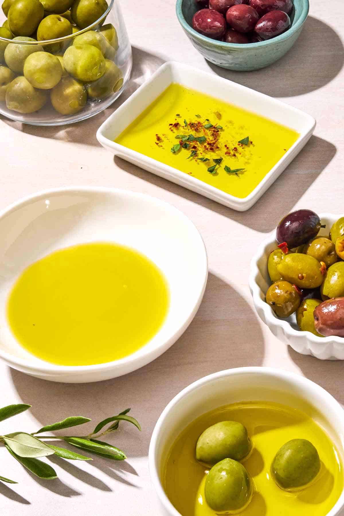 A bowl of olive oil surrounded by a clear bowl of green olives, a small bowl of kalamata olives, another bowl of olive oil seasoned with spices and herbs, a small bowl of mixed olives, and a small bowl with 3 green olives in olive oil.