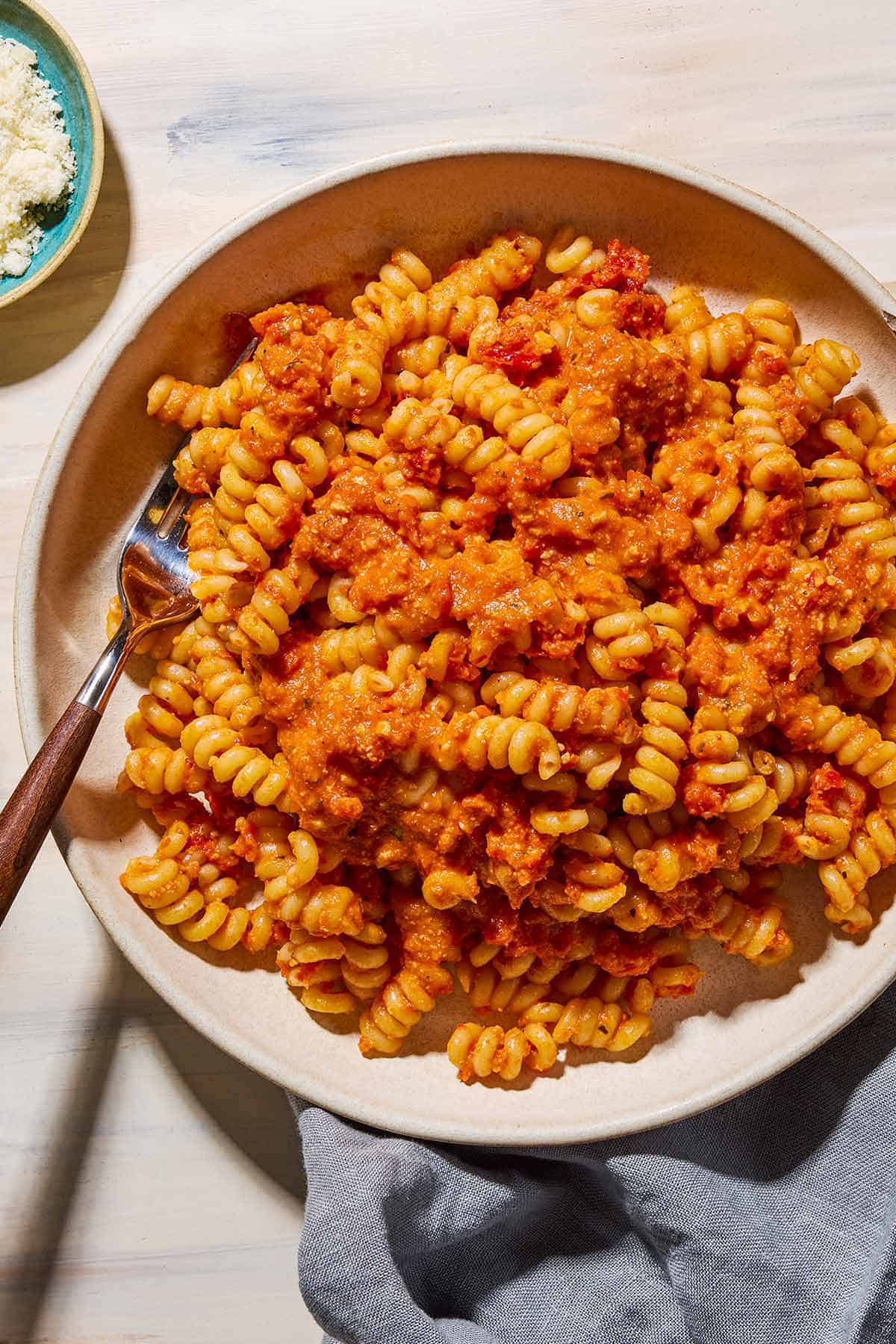 An overhead photo of sun dried tomato pesto pasta in a bowl with a fork. Next to this is a cloth napkin, and a bowl of grated cheese.