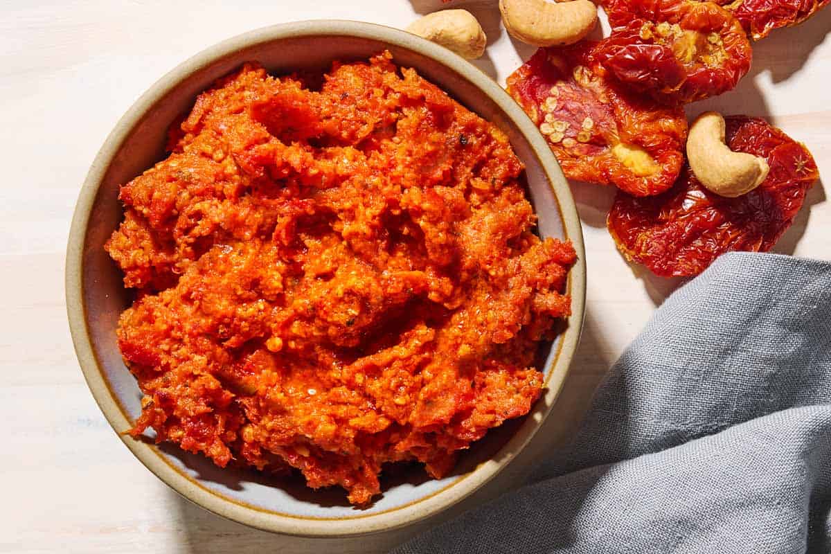 An overhead photo of sun dried tomato pesto in a bowl. Next to this is a cloth napkin, and some cashews and sun dried tomatoes.