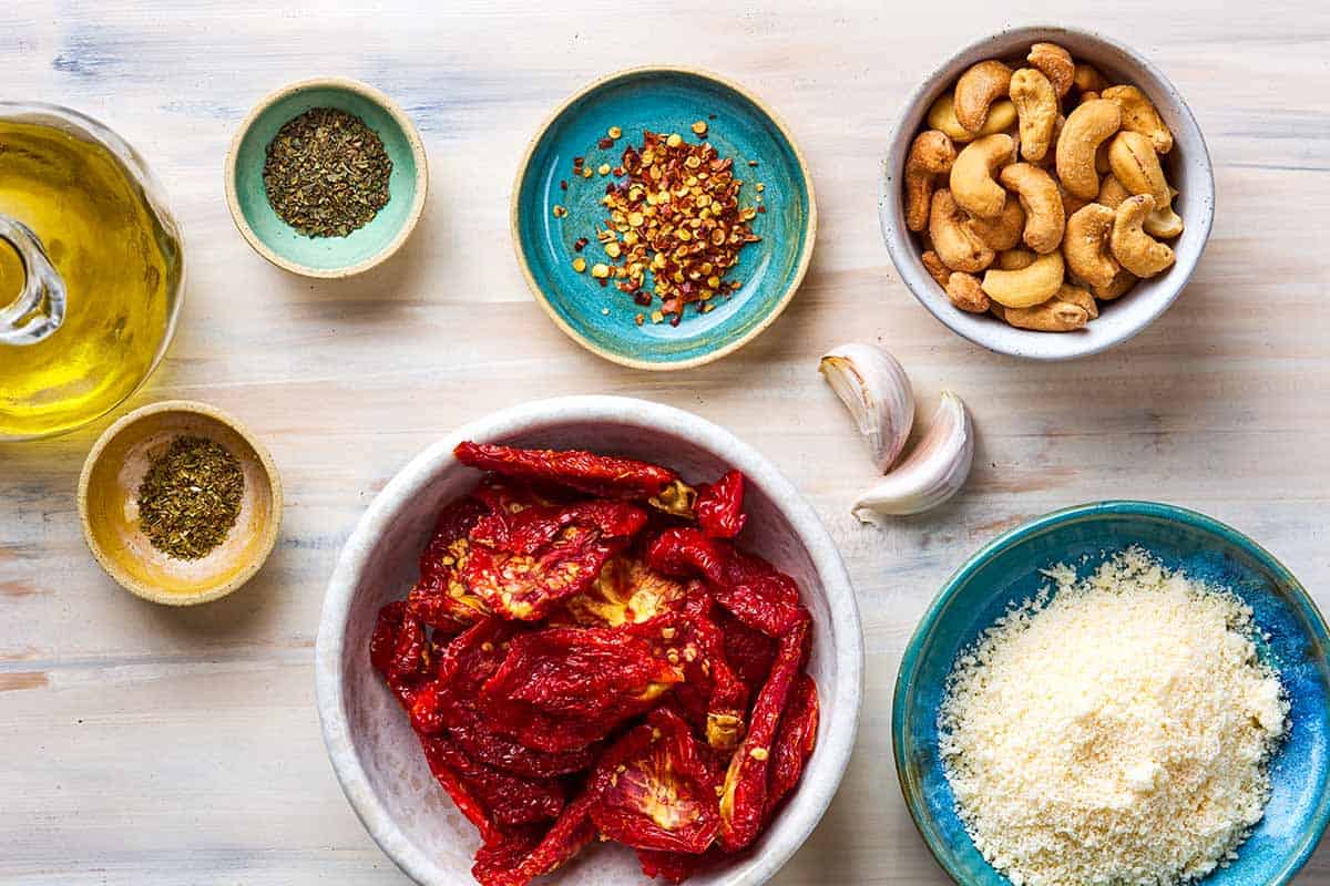 Ingredients for sun dried tomato pesto including sun dried tomatoes, grated parmesan cheese, cashews, garlic, oregano, basil, red pepper flakes, olive oil.