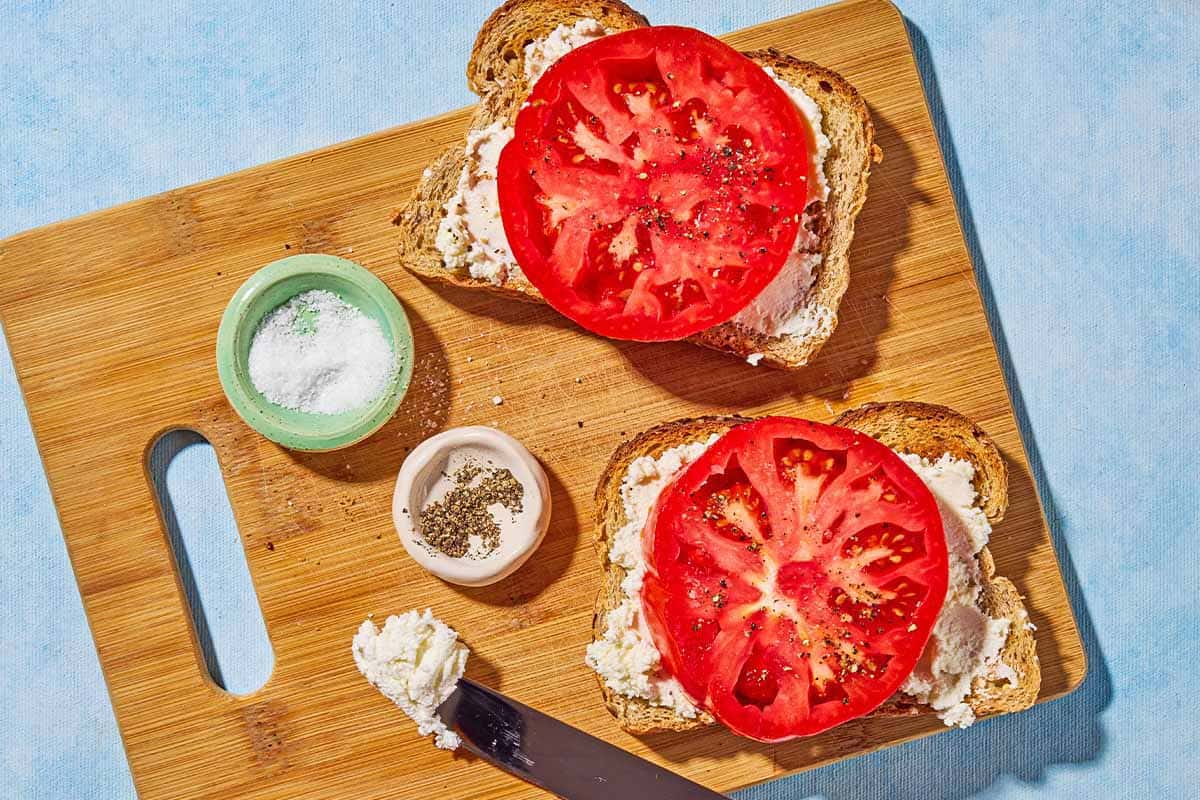 An overhead photo of two slices of toast topped with labneh, a tomato and salt and pepper next to bowls of salt and pepper and a knife on a cutting board.