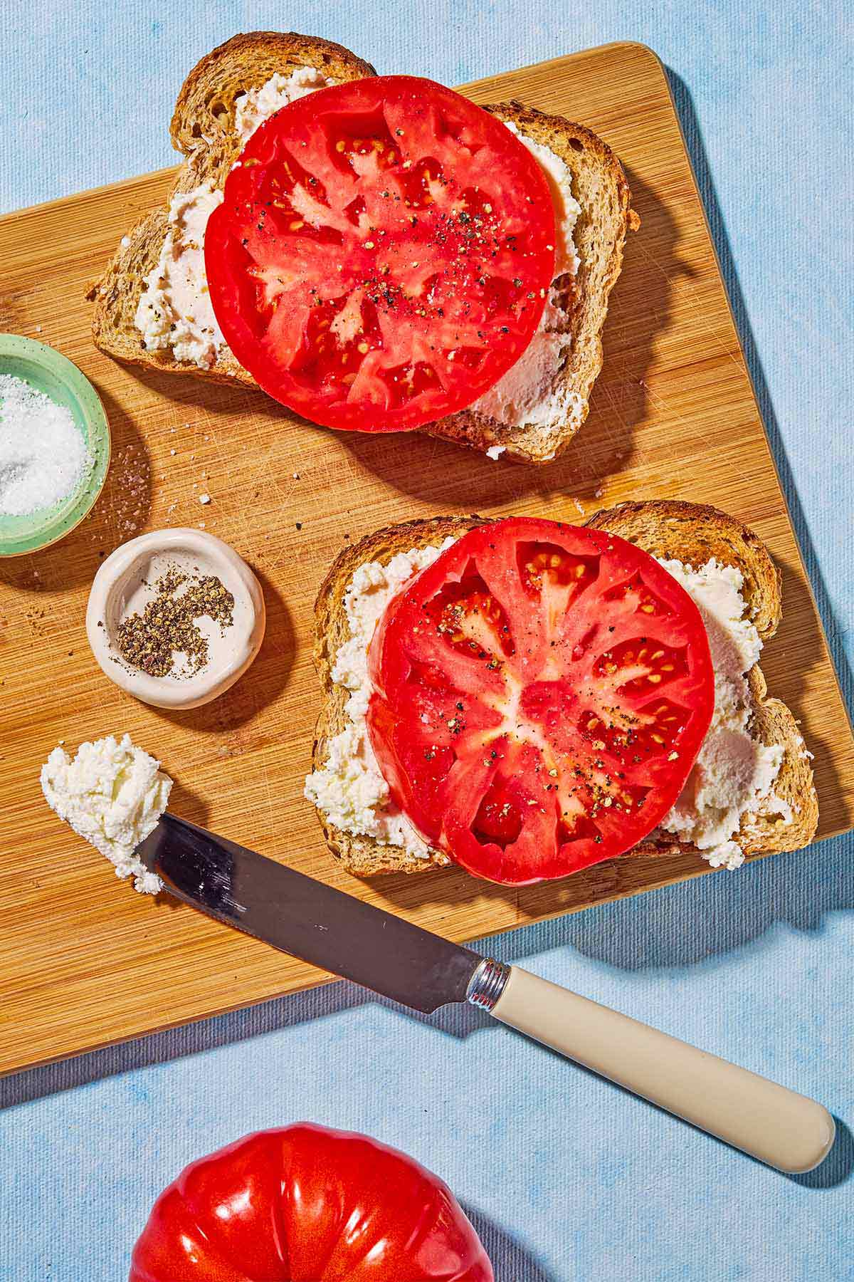 An overhead photo of two slices of toast topped with labneh, a tomato and salt and pepper next to bowls of salt and pepper and a knife on a cutting board.