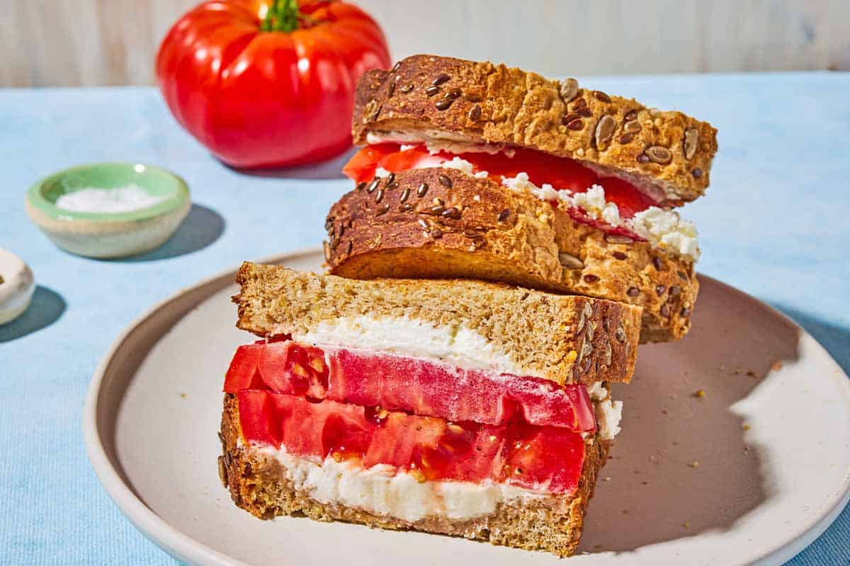 A close up of 2 tomato sandwiches stacked on top of each other on a plate. In the background is an heirloom tomato and a bowl of salt.