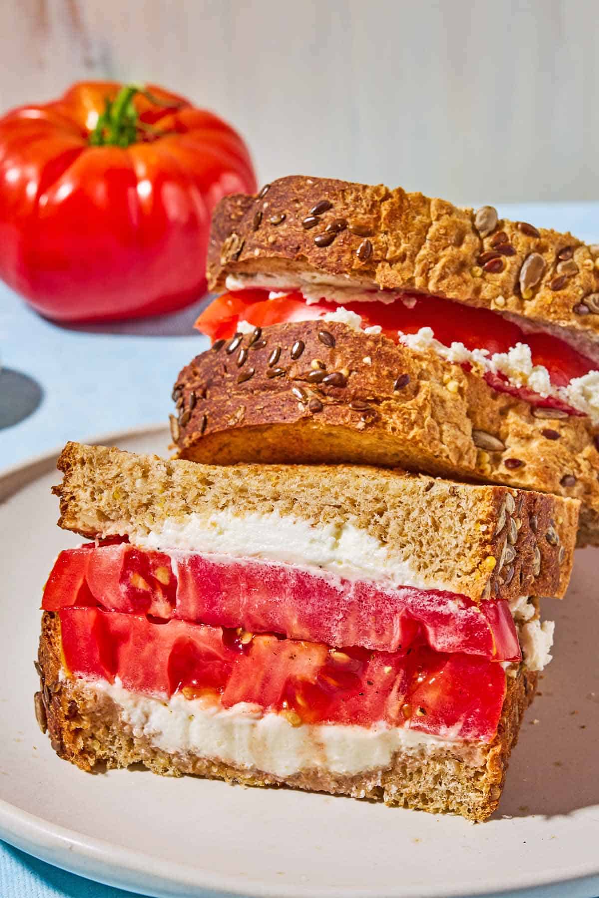 A close up of 2 tomato sandwiches stacked on top of each other on a plate. In the background is an heirloom tomato.