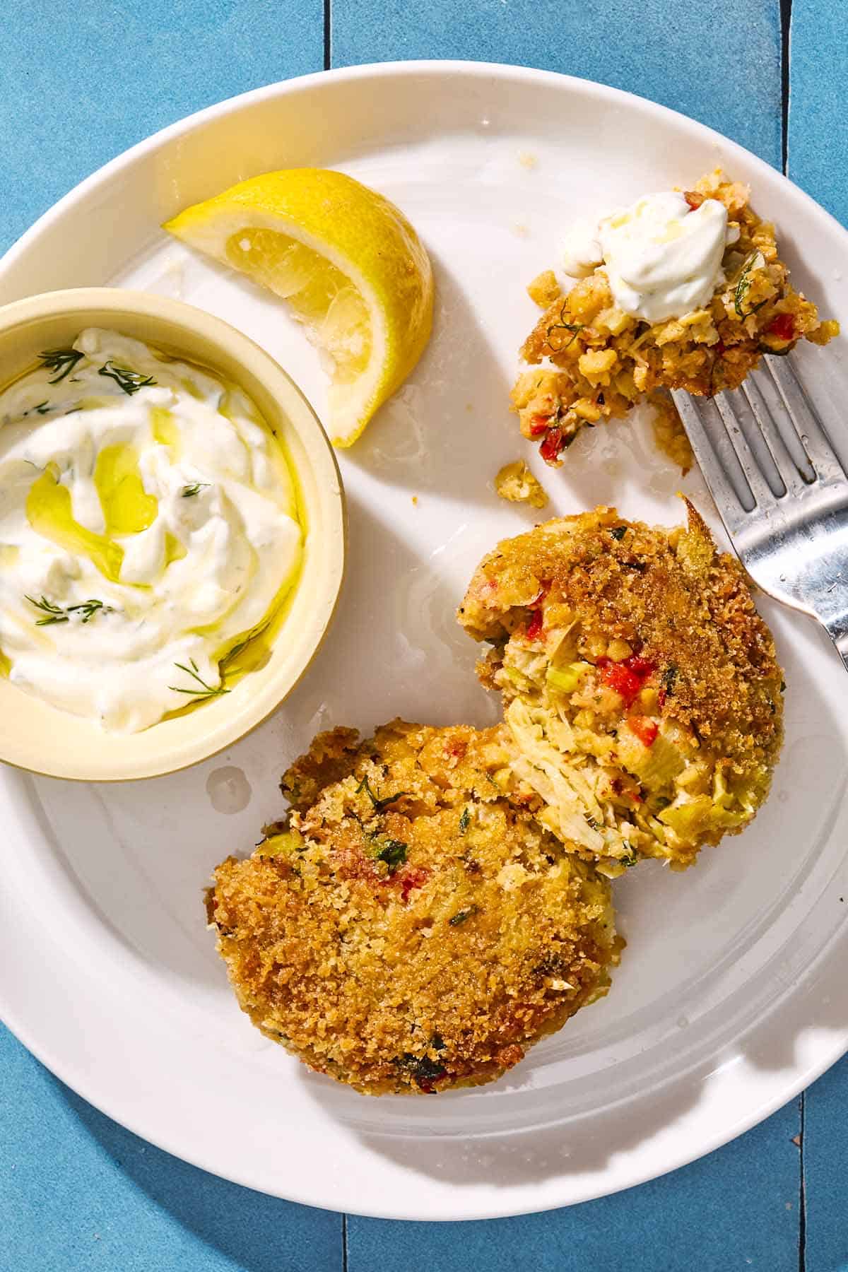 An overhead photo of 2 vegetarian crab cakes on a plate with a fork, a lemon wedge and a small bowl of tzatziki sauce.