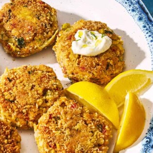 A closeup of vegetarian crab cakes on a serving platter with lemon wedges. One cake as a small dollop of tzatziki on it.