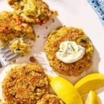 A closeup of vegetarian crab cakes on a serving platter with lemon wedges and a fork. One of the cakes has a small dollop of tzatziki on it.