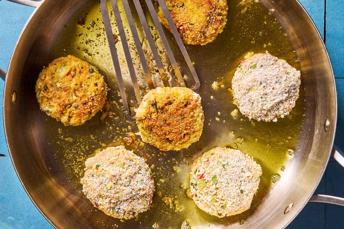 A closeup of 6 vegetarian crab cakes being fried in a skillet with spatula.