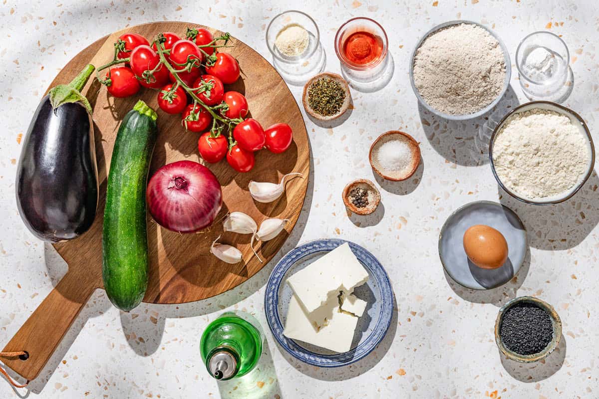 Ingredients for a savory galette including eggplant, zucchini, red onion, garlic, cherry tomatoes, salt, olive oil, za'atar, red wine vinegar, onion powder, black pepper, feta, parsley, whole wheat flour, all purpose flour, baking powder, olive oil, nigella seeds and an egg.