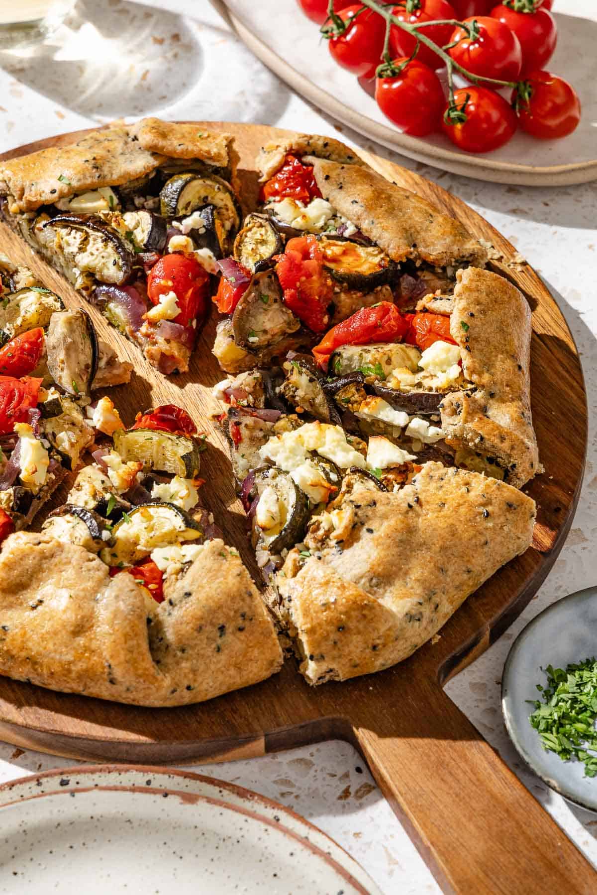 A close up of a savory galette cut into slices on a wooden serving board. Next to this is a plate of cherry tomatoes, a bowl of minced parsley and a stack of plates.