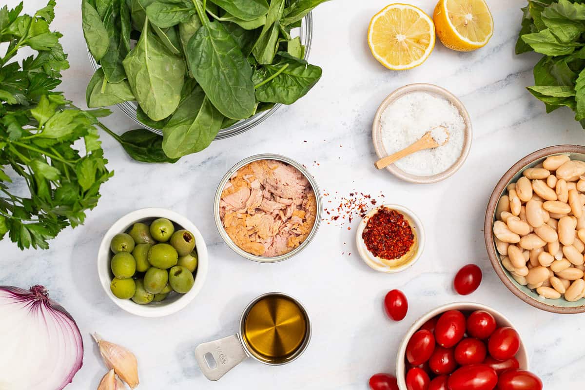 Ingredients for tuna white bean salad including canned tuna, cannellini beans, lemons, garlic, olive oil, aleppo pepper, kosher salt, cherry tomatoes, red onion, parsley, green olives, basil and baby spinach.