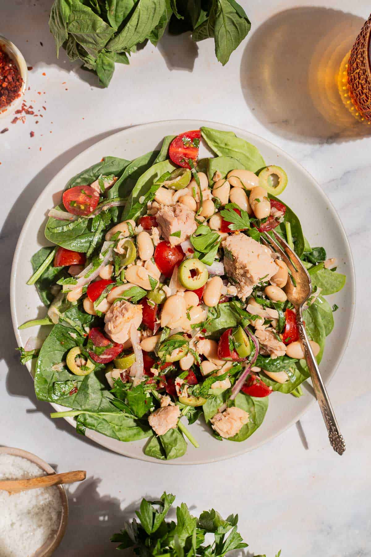 An overhead photo of a serving tuna white bean salad on a plate with a fork. Next to this is a glass of water, sprigs of basil and parsley, and bowls of kosher salt and aleppo pepper.