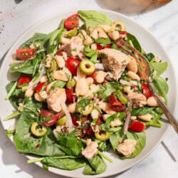 A close up photo of a serving tuna white bean salad on a plate with a fork. Next to this is a glass of water, sprigs of basil and parsley, and a bowl of kosher salt.