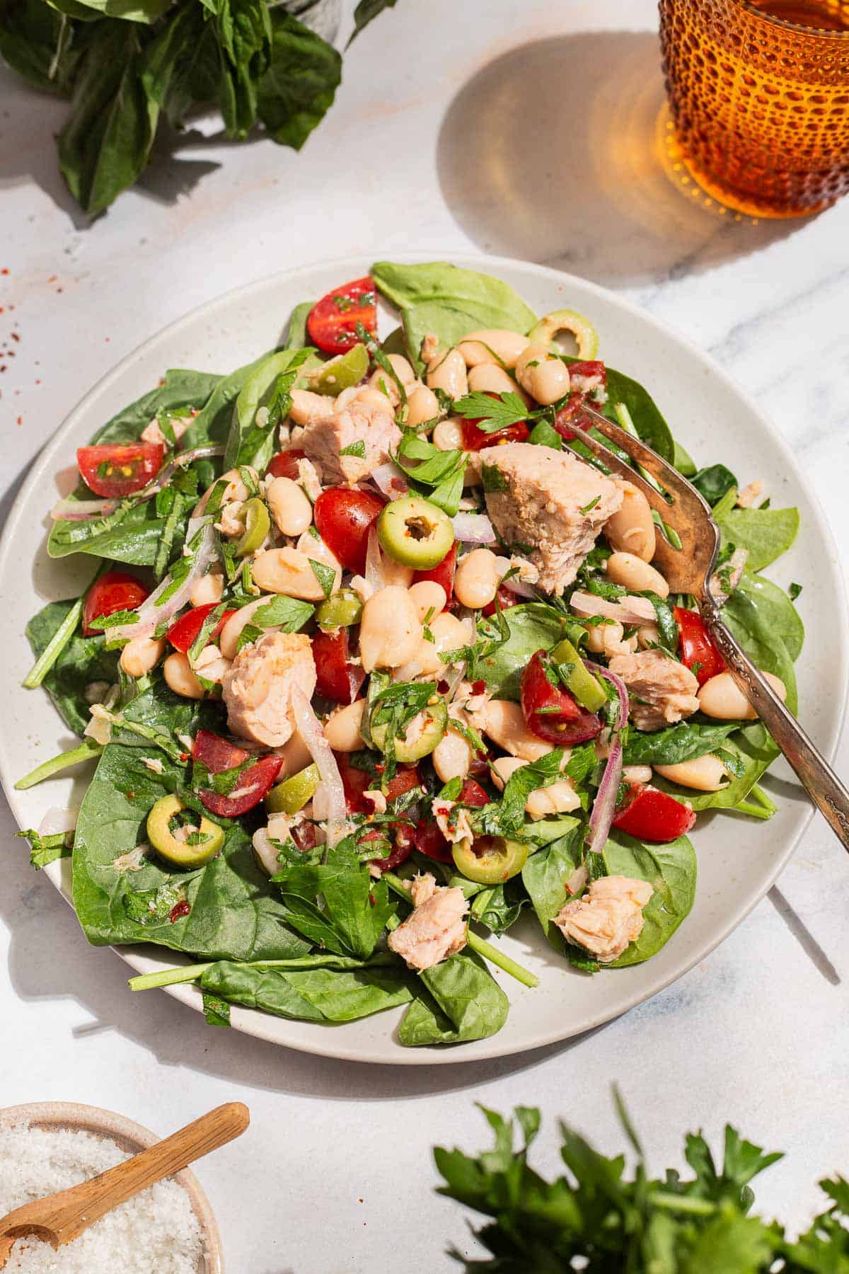 A close up photo of a serving tuna white bean salad on a plate with a fork. Next to this is a glass of water, sprigs of basil and parsley, and a bowl of kosher salt.