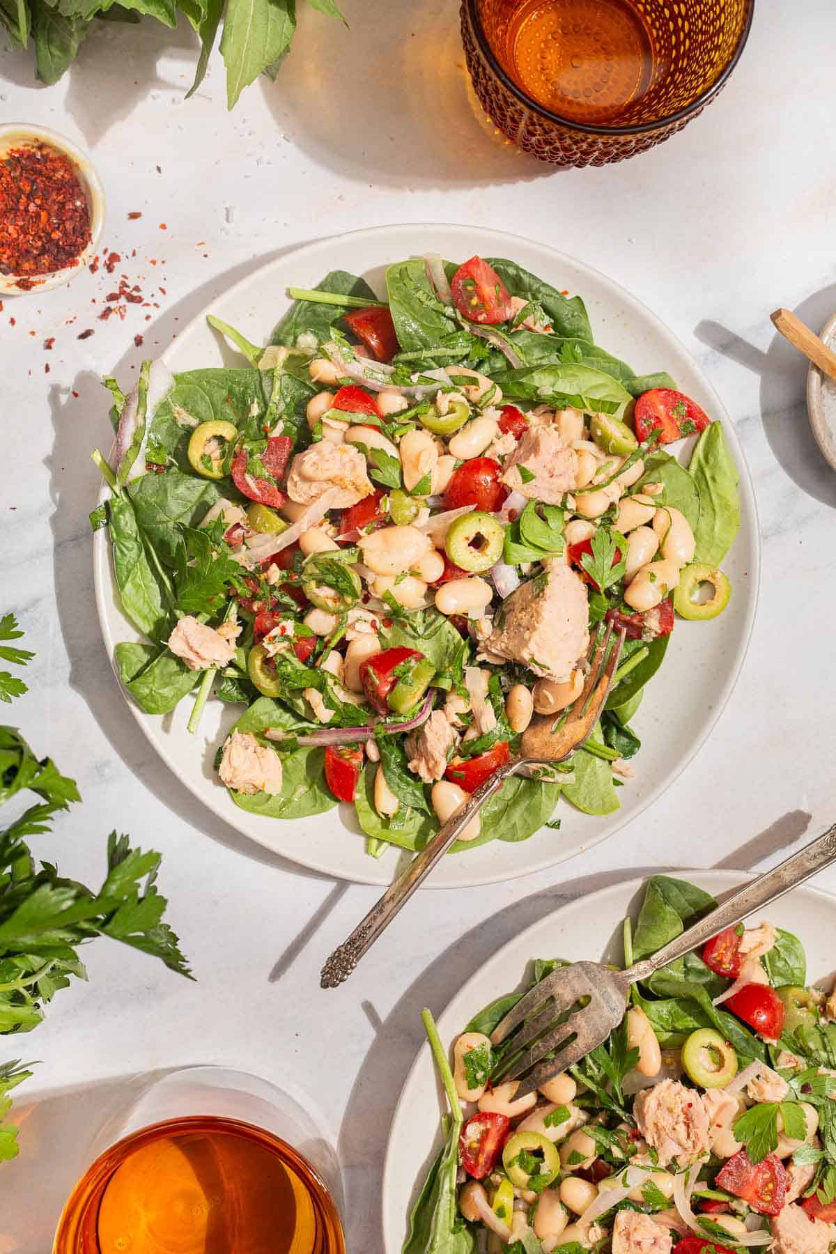 An overhead photo of a serving tuna white bean salad on a plate with a fork. Next to this is another serving of the salad on a plate with a fork, 2 glasses of water, sprigs of basil and parsley, and a bowl of aleppo pepper.