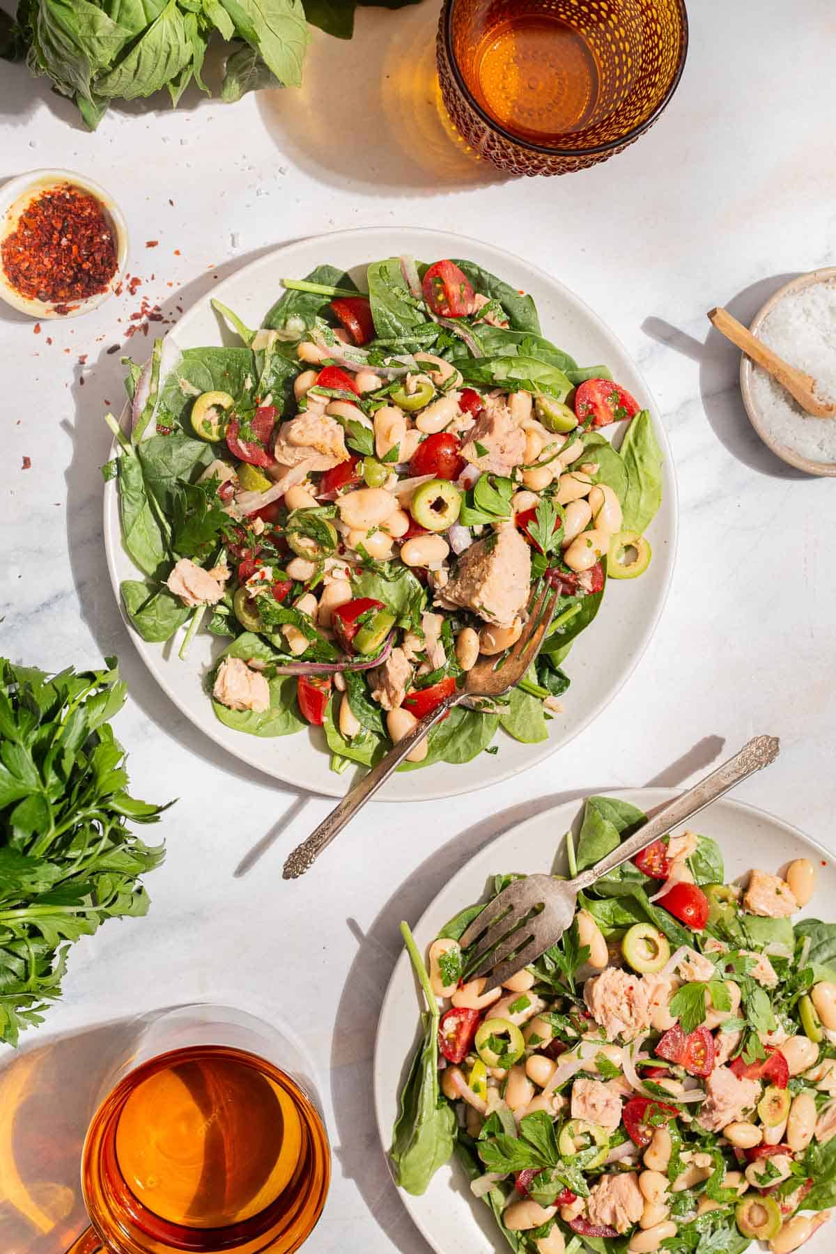 An overhead photo of a serving tuna white bean salad on a plate with a fork. Next to this is another serving of the salad on a plate with a fork, 2 glasses of water, sprigs of basil and parsley, and bowls of kosher salt and aleppo pepper.