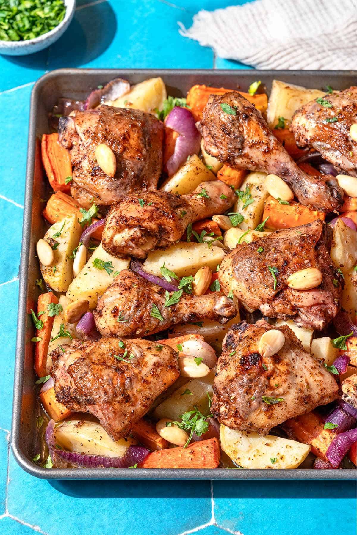 A close up photo of sheet pan za'atar chicken with veggies next to a small bowl of chopped parsley and a kitchen towel.