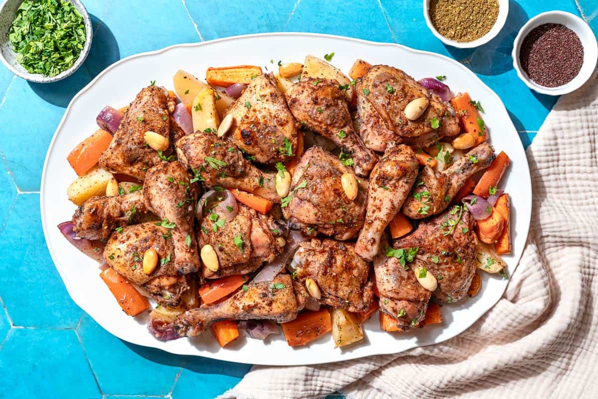 An overhead photo of sheet pan za'atar chicken with veggies on a platter next to a kitchen towel and small bowls of chopped parsley, za'atar and sumac.