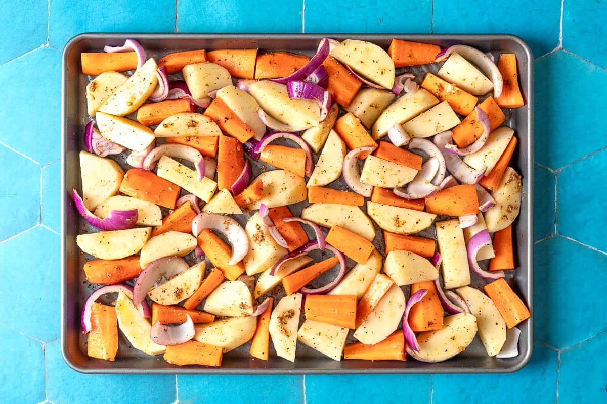 An overhead photo of seasoned, uncooked potatoes, carrots and red onions on a sheet pan.