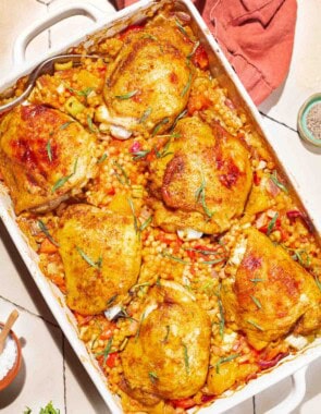 An overhead photo of the one pan tomato chicken with baharat, tarragon, and couscous in a baking dish. Next to this is a cloth napkin, small bowls of salt, pepper and sprig of tarragon.