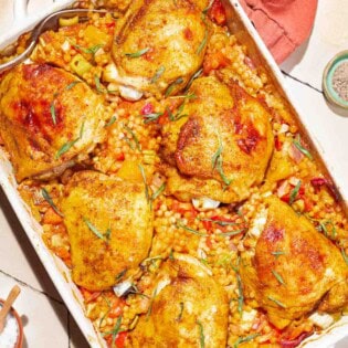 An overhead photo of the one pan tomato chicken with baharat, tarragon, and couscous in a baking dish. Next to this is a cloth napkin, small bowls of salt, pepper and sprig of tarragon.