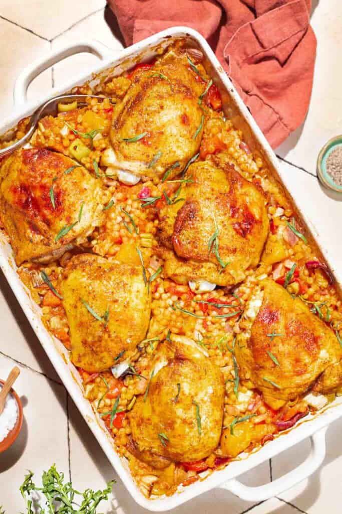 An overhead photo of the one pan tomato chicken with baharat, tarragon, and couscous in a baking dish. Next to this is a cloth napkin, small bowls of salt, pepper and sprig of tarragon.