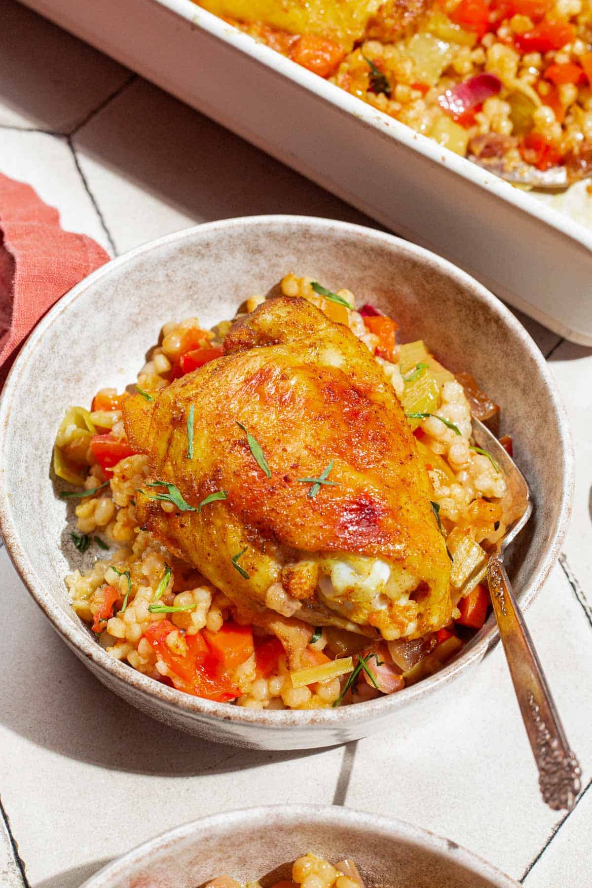 A close up of a serving of the one pan tomato chicken with baharat, tarragon, and couscous in a bowl with a fork. Behind this is the baking dish with the rest of the tomato chicken.