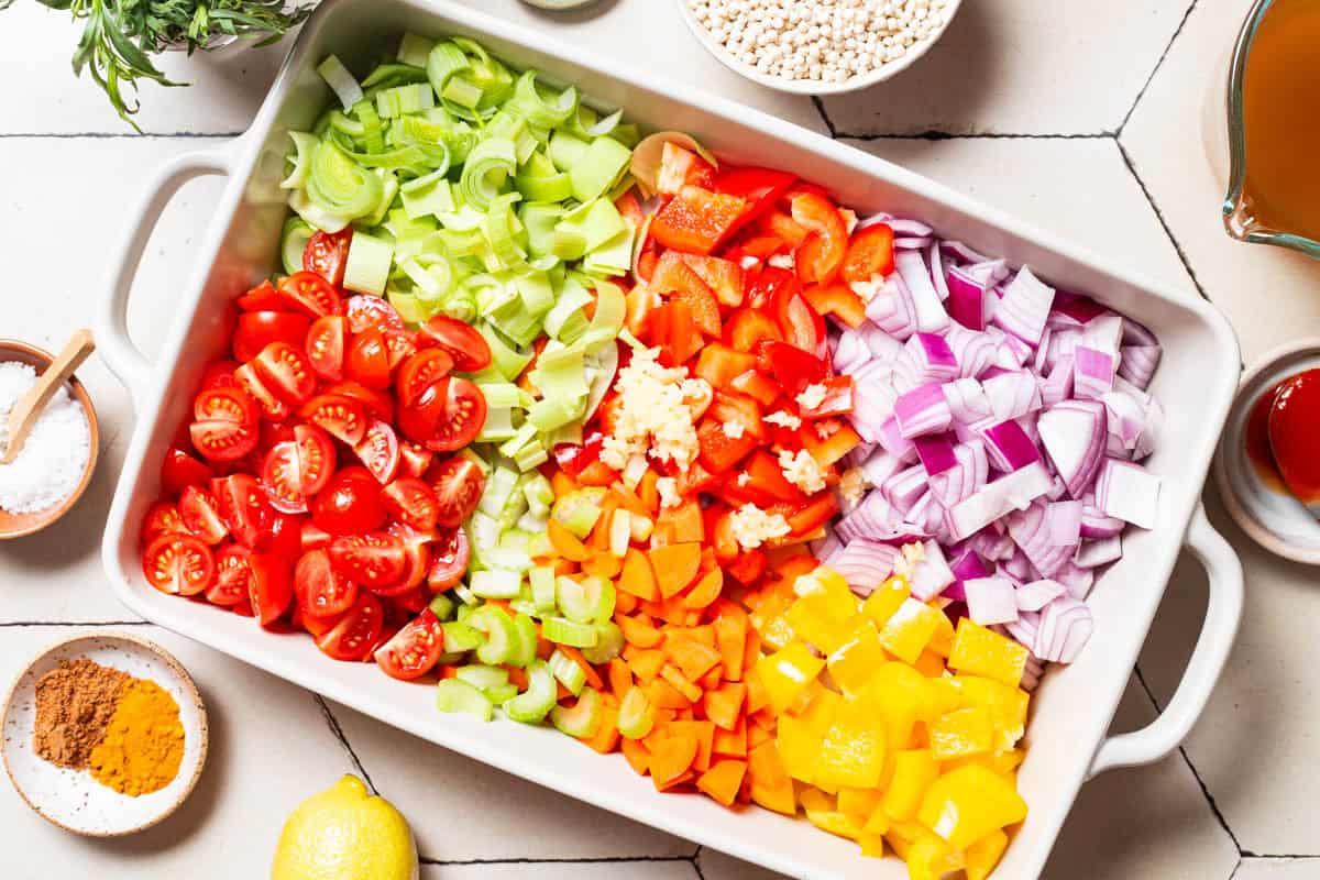 An overhead photo of the chopped vegetables for the one pan tomato chicken in a baking dish just before being mixed together. Next to this is a bunch of tarragon, a bowl of pearl couscous, broth, tomato paste, a lemon, a bowl of baharat and turmeric, and a bowl of salt.