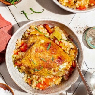 An overhead photo of a serving of the one pan tomato chicken with baharat, tarragon, and couscous in a bowl with a fork. Next to this is a lemon half, small bowls of salt, pepper and tarragon, and a bowl with another serving of the chicken.