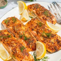 A close up of 4 baked dijon salmon fillets on a serving platter with lemon wedges and a fork. Next to this is a bowl of black pepper and a bunch of dill.