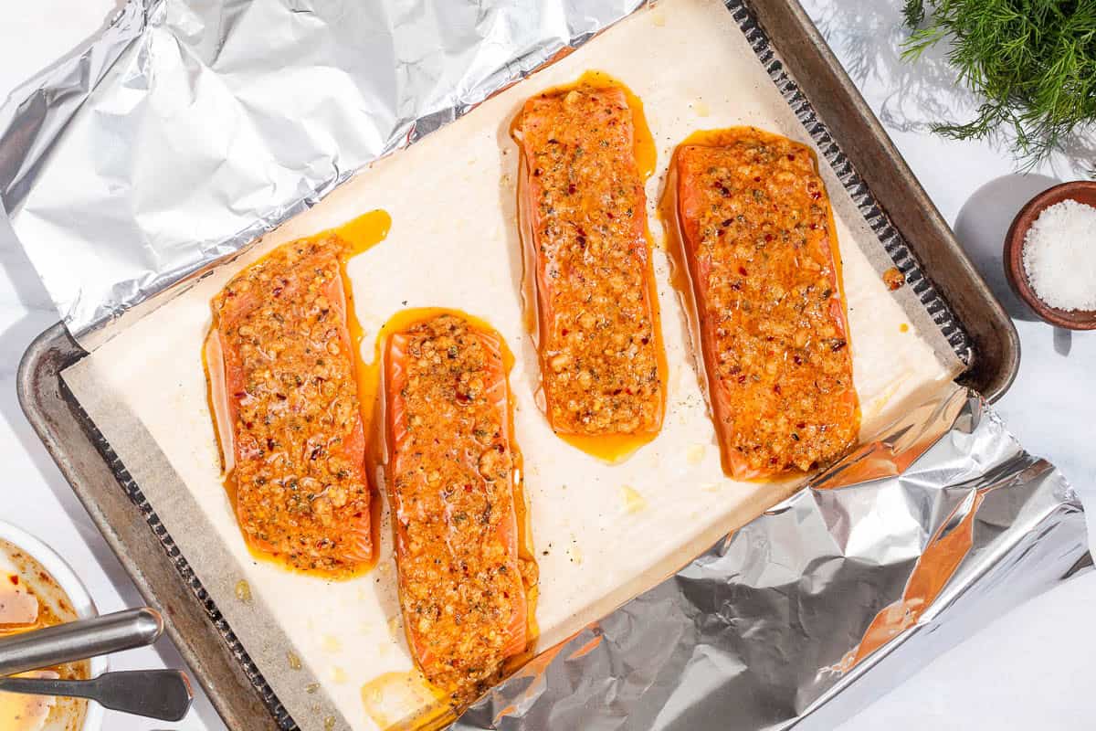 An overhead photo of 4 unbaked dijon salmon fillets on a parchment and foil lined baking sheet. Next to this is a bunch of dill and a bowl of salt.