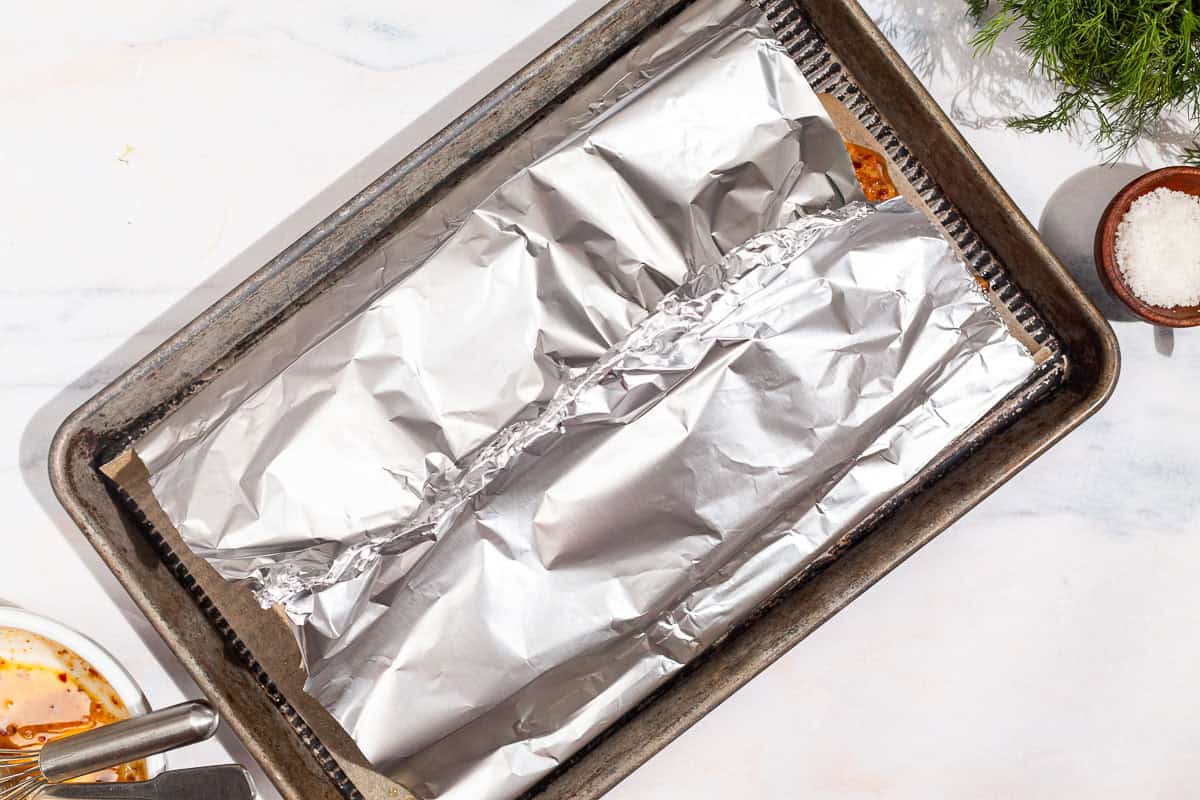 An overhead photo of unbaked dijon salmon fillets on a piece of parchment, wrapped in foil on a baking sheet. Next to this is a bowl of salt and some dill.