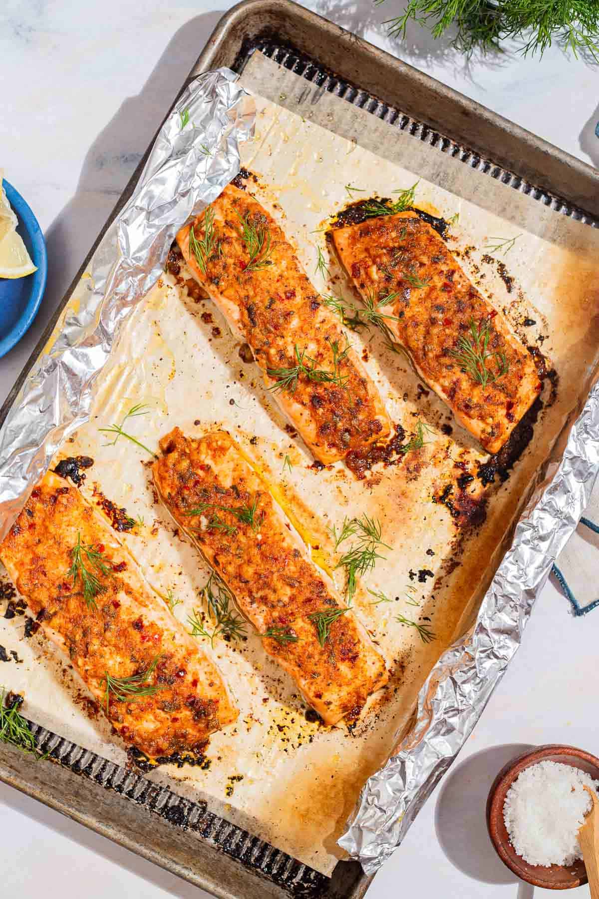 An overhead photo of 4 baked dijon salmon fillets on a parchment and foil lined baking sheet. Next to this is a bowl of salt.