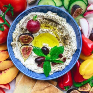 An overhead photo of homemade labneh in a bowl, garnished with herbs, olives, figs, a radish and a drizzle of olive oil surrounded by various fruits and raw vegetables and a bowl of za'atar.