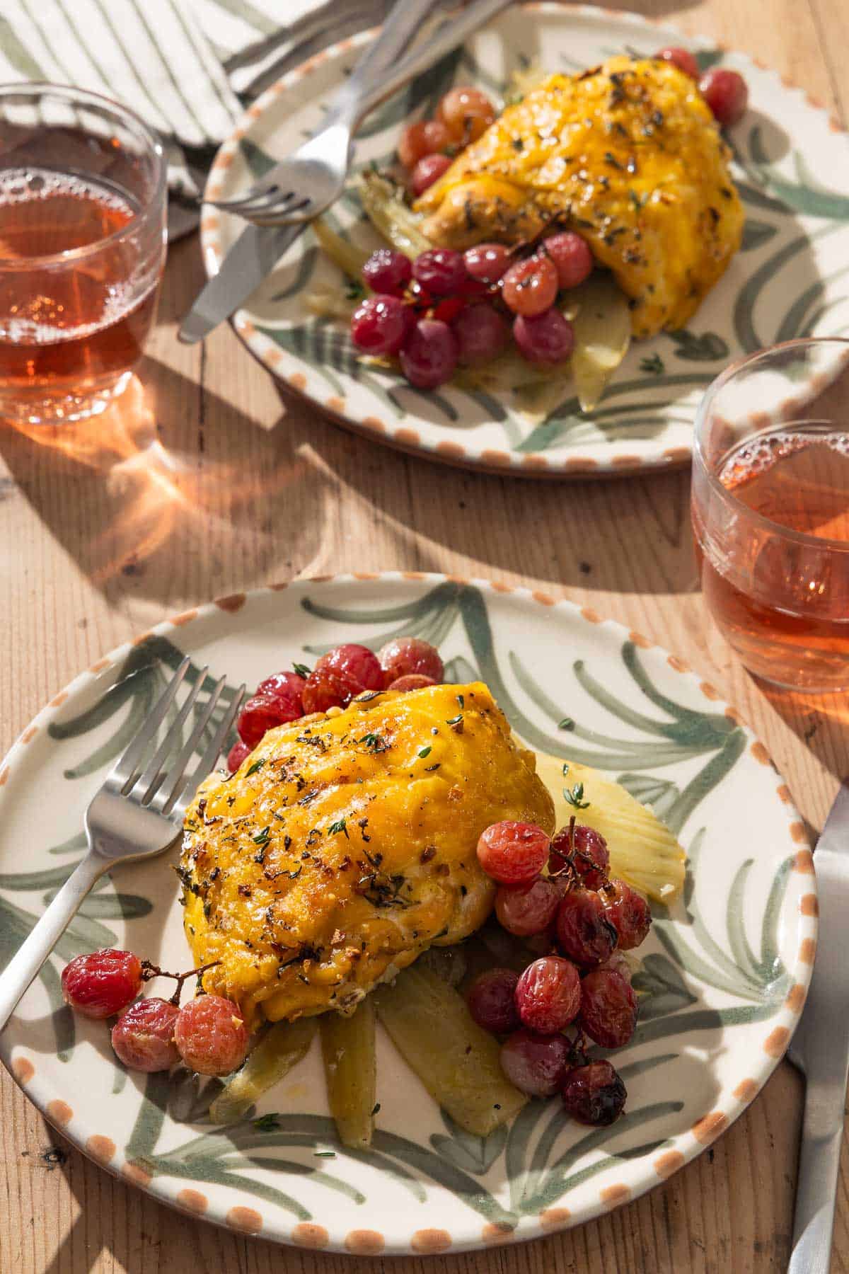 2 braised chicken thighs, each on its own plate with grapes, fennel and utensils. Next to these are 2 glasses of rosé.