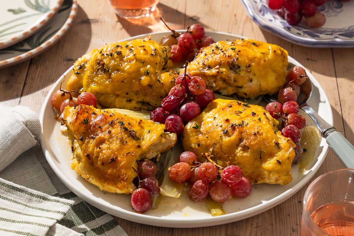 A close up of 4 braised chicken thighs with grapes and fennel on a serving platter with a spoon. Next to this is a cloth napkin, a stack of 2 plates, 2 glasses of rosé, and a plate of red grapes.