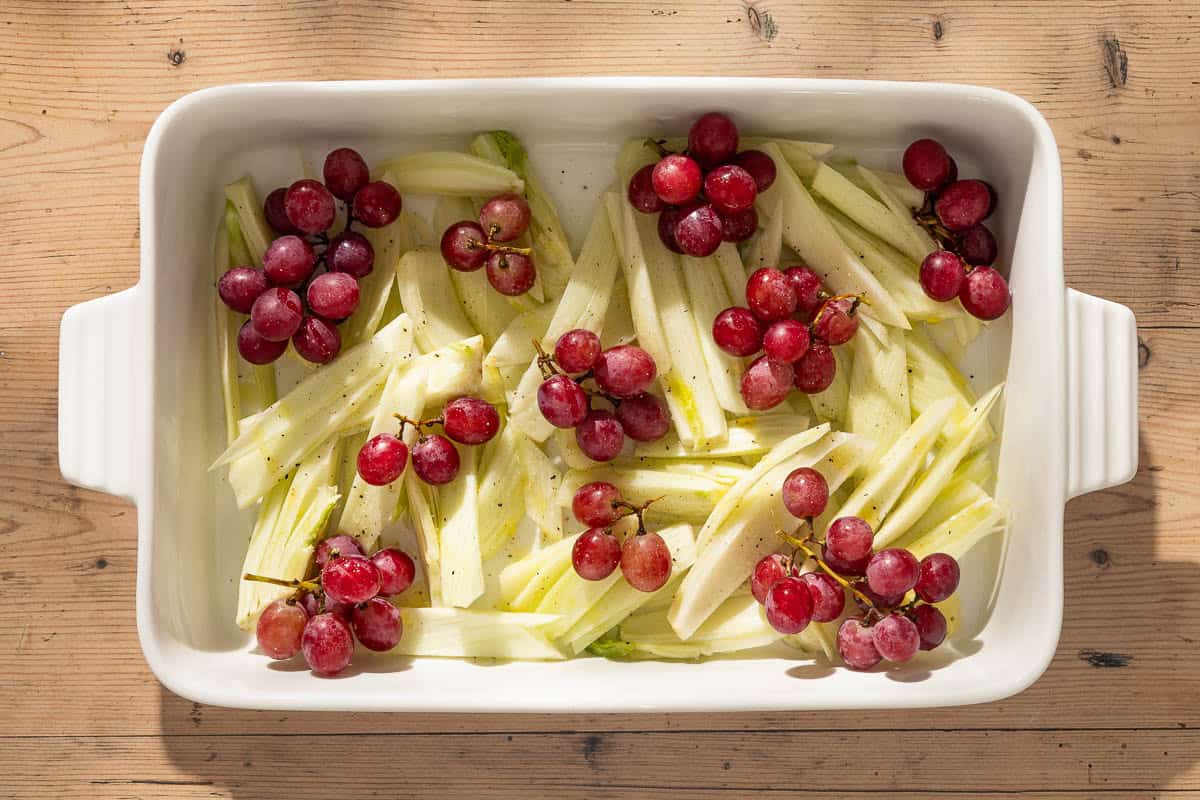 An overhead photo of uncooked red grapes and fennel drizzled with olive oil and seasoned with salt and black pepper in a baking dish.